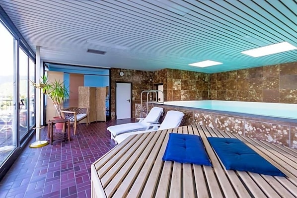 Zona de piscina interior con luz solar, sillones de descanso y una terraza de madera.