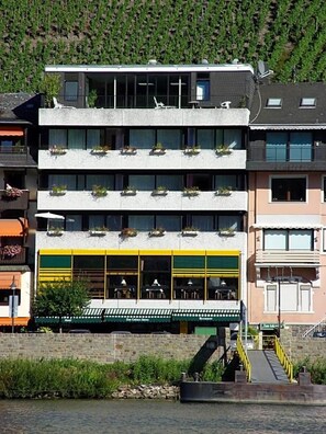 Un hotel de varios pisos con balcones, un restaurante con terraza y vistas al río.
