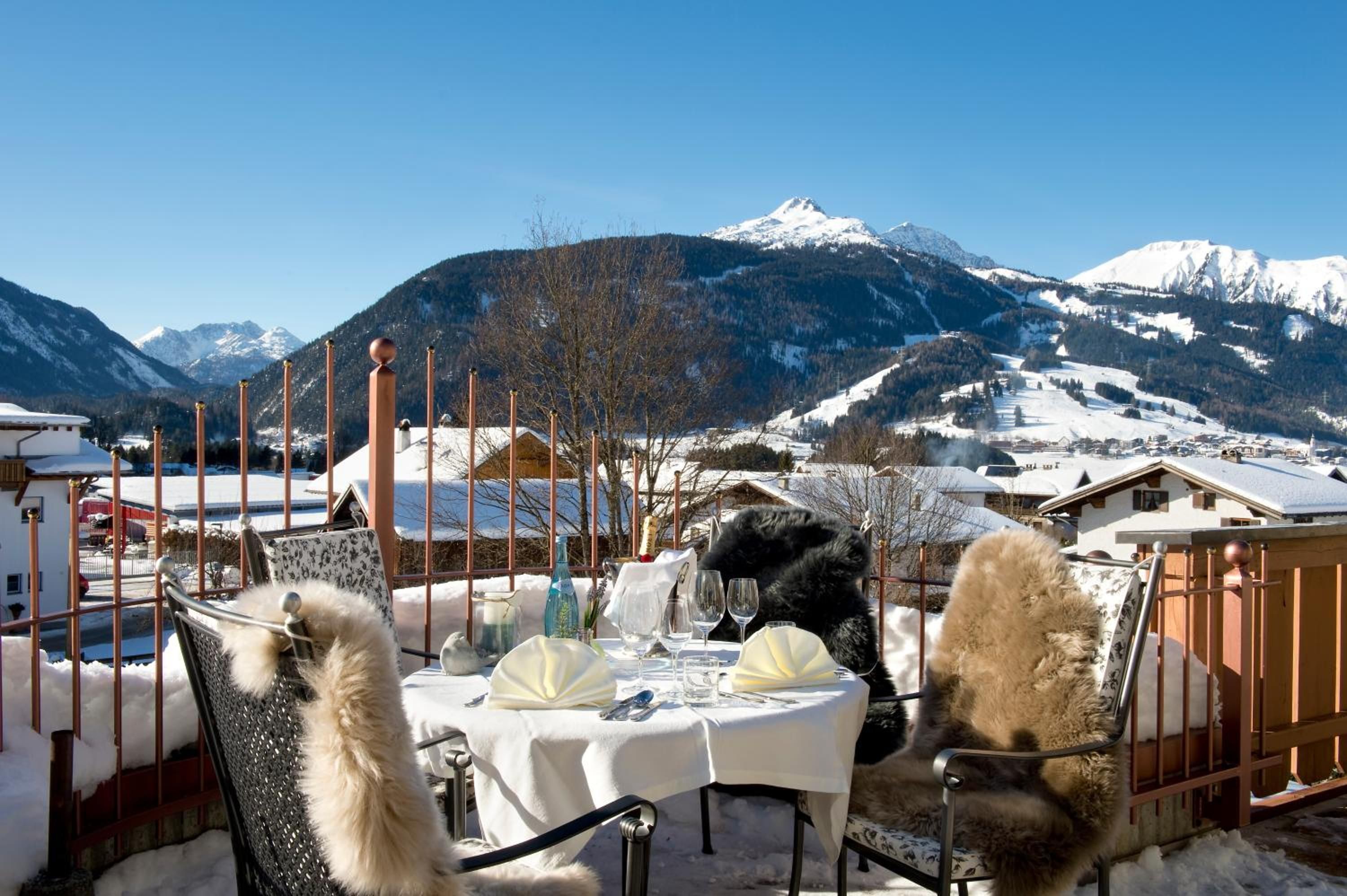 A table set outdoors with a mountain view, a chair with a fur throw, and a metal fence.