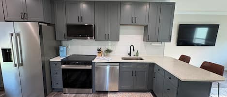 Una cocina moderna con armarios grises, electrodomésticos de acero inoxidable, un televisor de pantalla plana y suelo de madera.