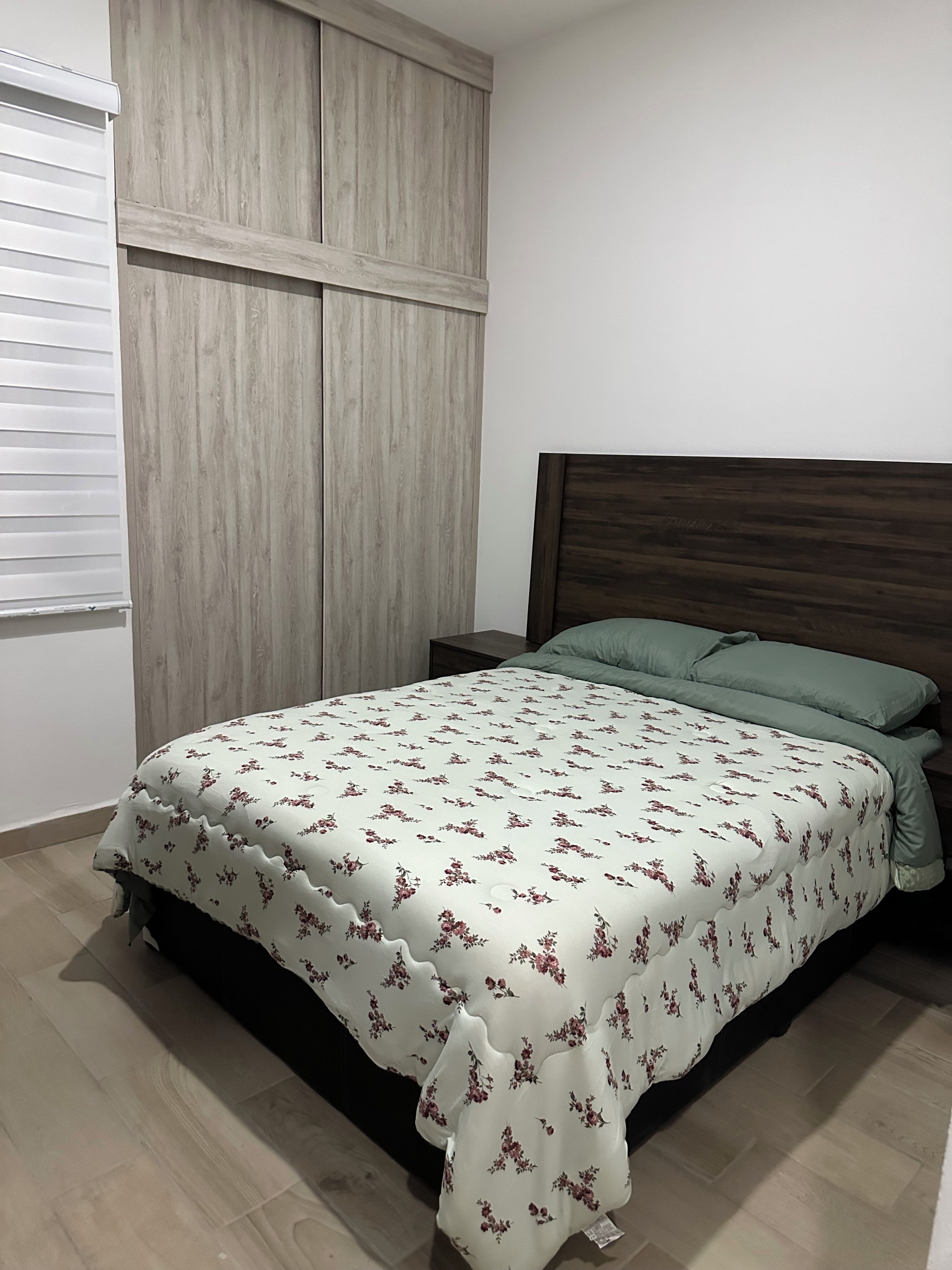 A bedroom with a bed, a nightstand, a wooden headboard, and a window with blinds.