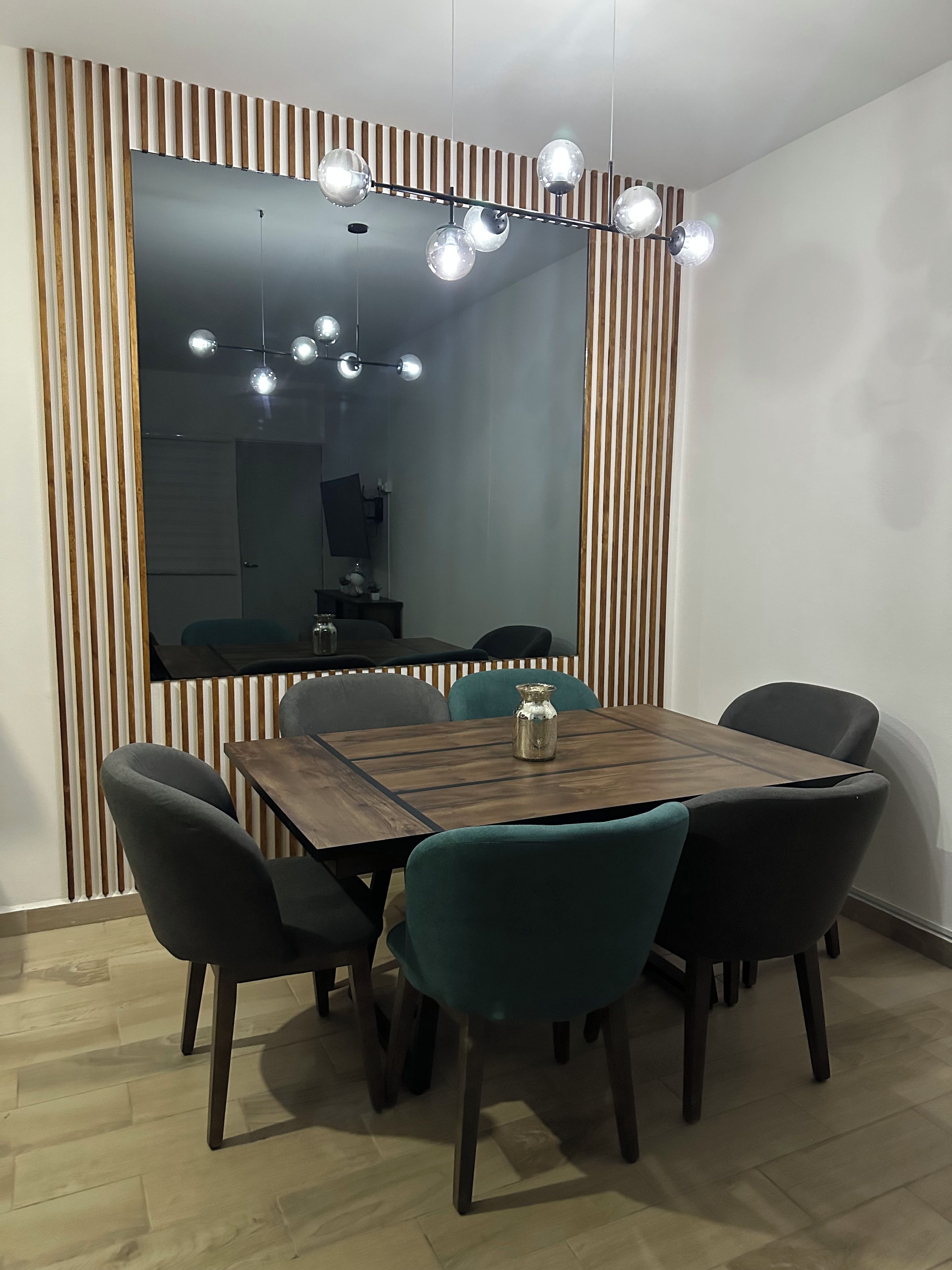Un comedor con una mesa de madera, cuatro sillas, un espejo grande y una lámpara moderna.