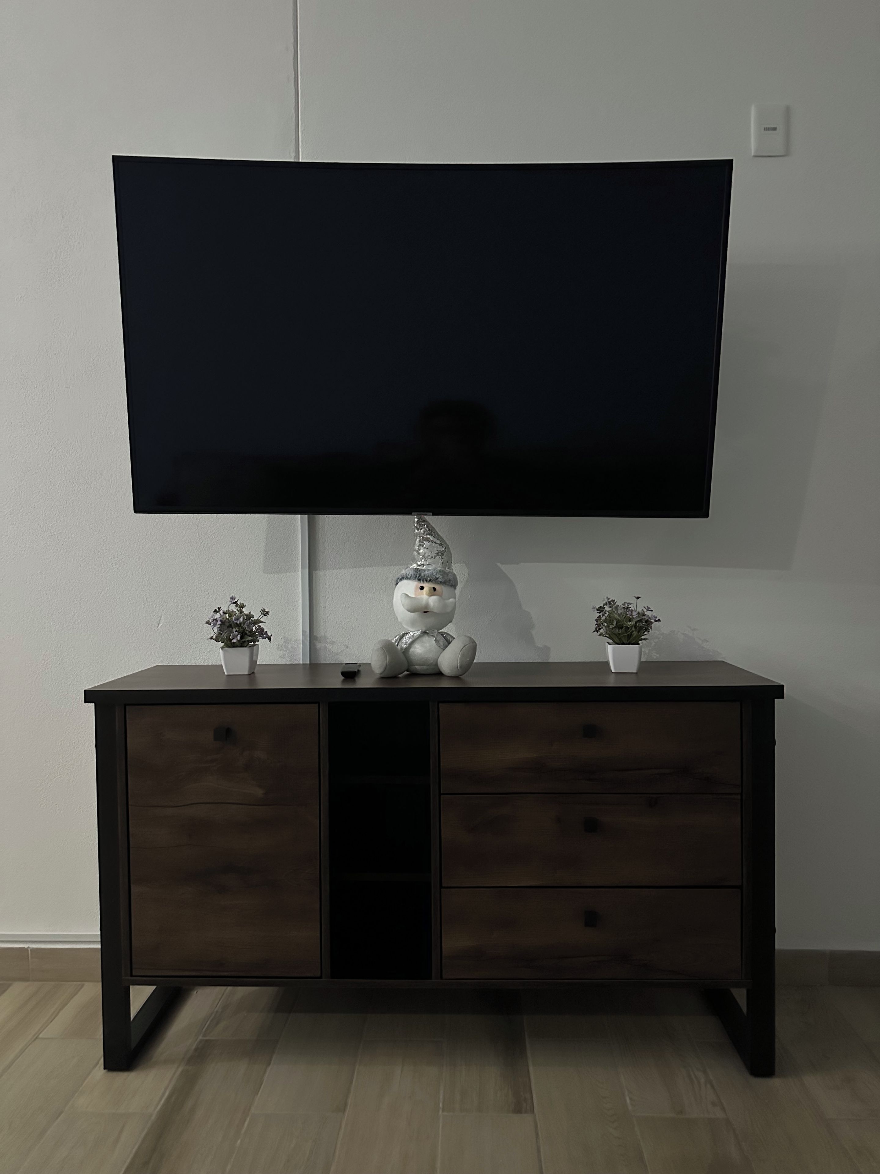 A flat-screen TV mounted on a wall above a dark wooden cabinet with two potted plants and a decorative item.