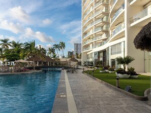 Une piscine entourée de chaises longues, de palmiers et d’un bâtiment moderne doté de balcons.