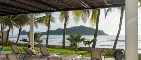 A covered outdoor seating area with lounge chairs and a view of the sea and mountains.