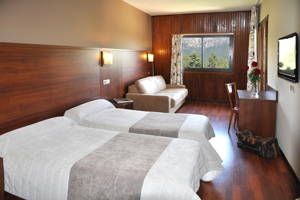 A hotel room with two beds, a wooden headboard, a window with a view, a desk with a chair, and a vase with flowers.