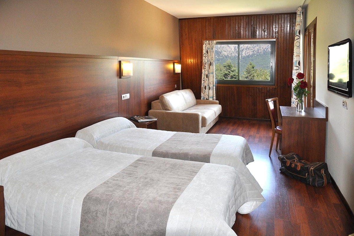 A hotel room with two beds, a wooden headboard, a window with a view, a desk with a chair, and a vase with flowers.