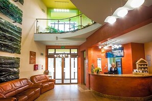 Zona de recepción de un hotel con un mostrador para recepcionistas, una escalera a un nivel superior y un mostrador de recepción con un cartel.