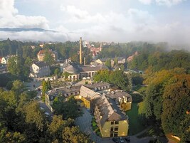 Un complejo histórico de edificios con varias estructuras, incluyendo una torre destacada, rodeado de vegetación exuberante y un paisaje montañoso.