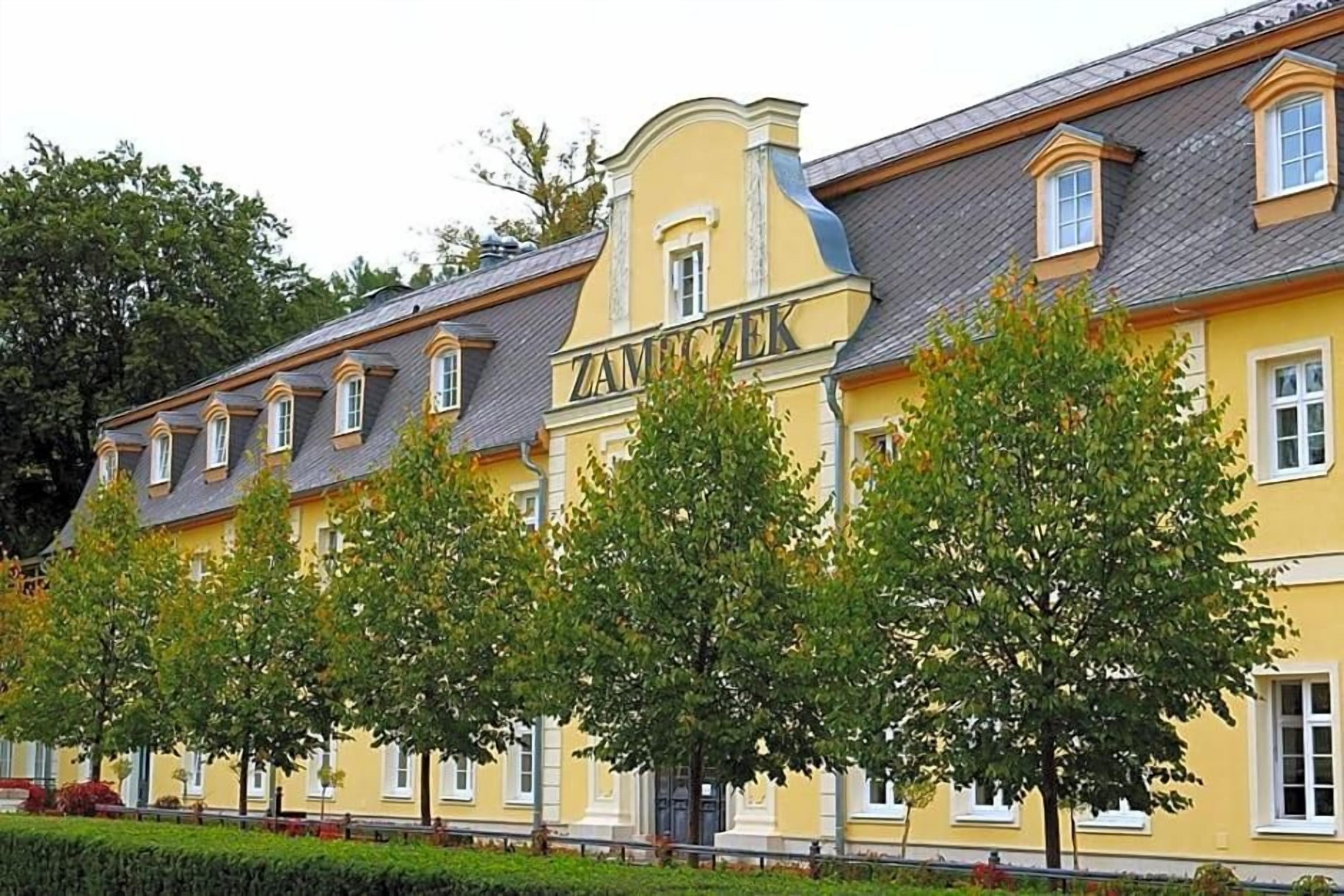 A yellow building with the name 'ZAMECZEK' on the facade, multiple windows, and trees in front.