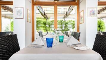 Ein Restaurant mit einem langen Esstisch, gerahmten Kunstwerken an den Wänden und Blick auf Grünfläche vor den Fenstern.
