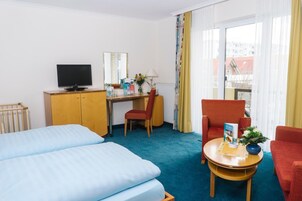 A hotel room with two single beds, a desk with a computer and a TV, a red armchair, a small table with a plant, and a window with curtains.