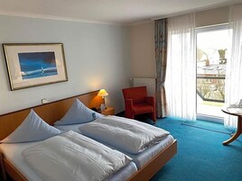 A hotel room with a bed, a chair, a table, a lamp, a framed picture, and a view of the outside.