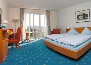A hotel room with a bed, a desk, a chair, a small table, and a view of the city through the window. - Aparthotel Kachelot (Borkum)