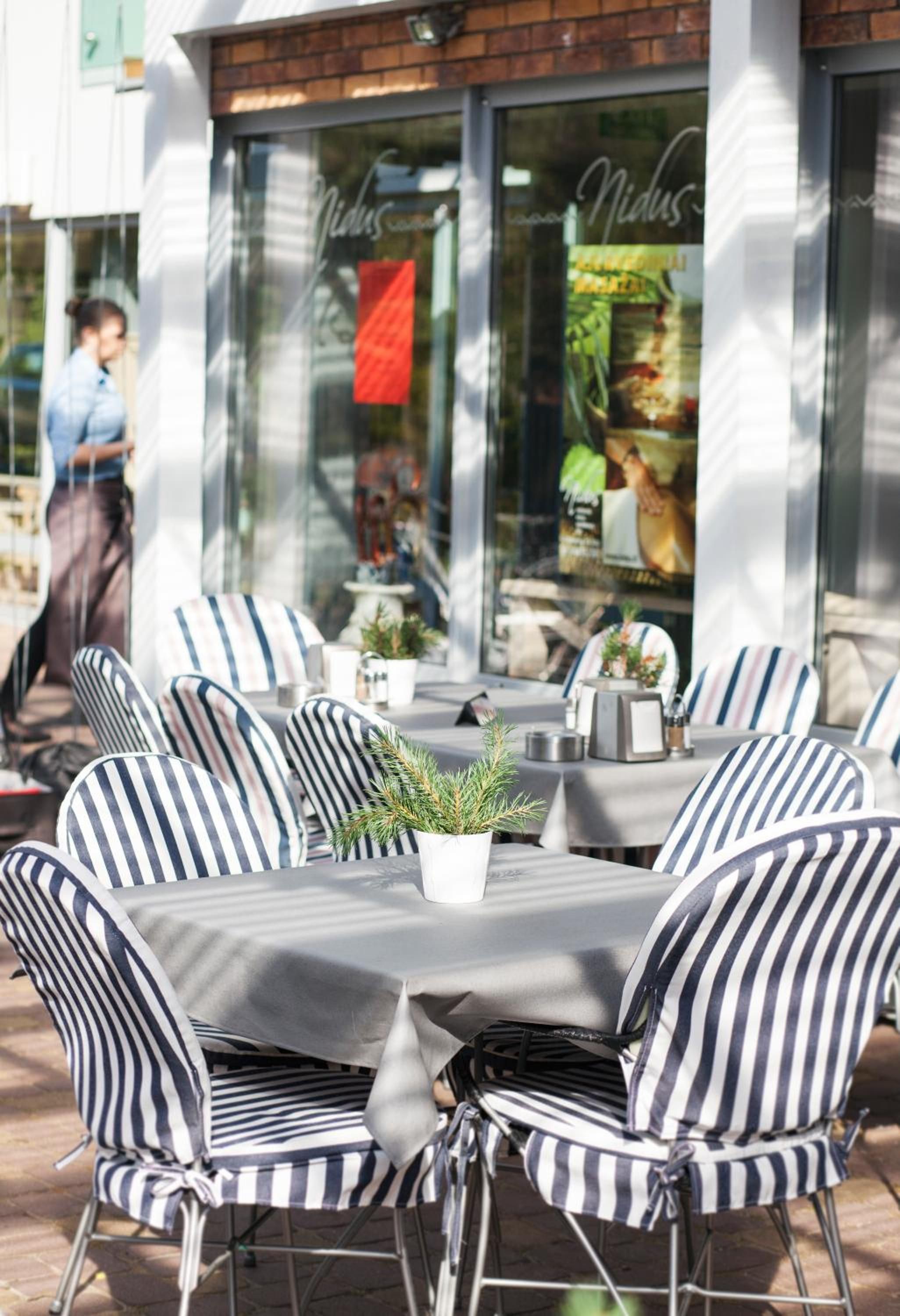 An outdoor seating area with striped chairs and tables, a potted plant, and a storefront with a red sign.