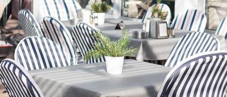 An outdoor seating area with striped chairs and tables, a potted plant, and a storefront with a red sign.