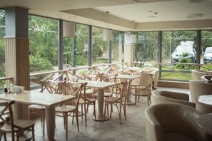 Een ruime binnenruimte met meerdere tafels en stoelen, grote ramen bieden uitzicht op groen en geparkeerde auto's.