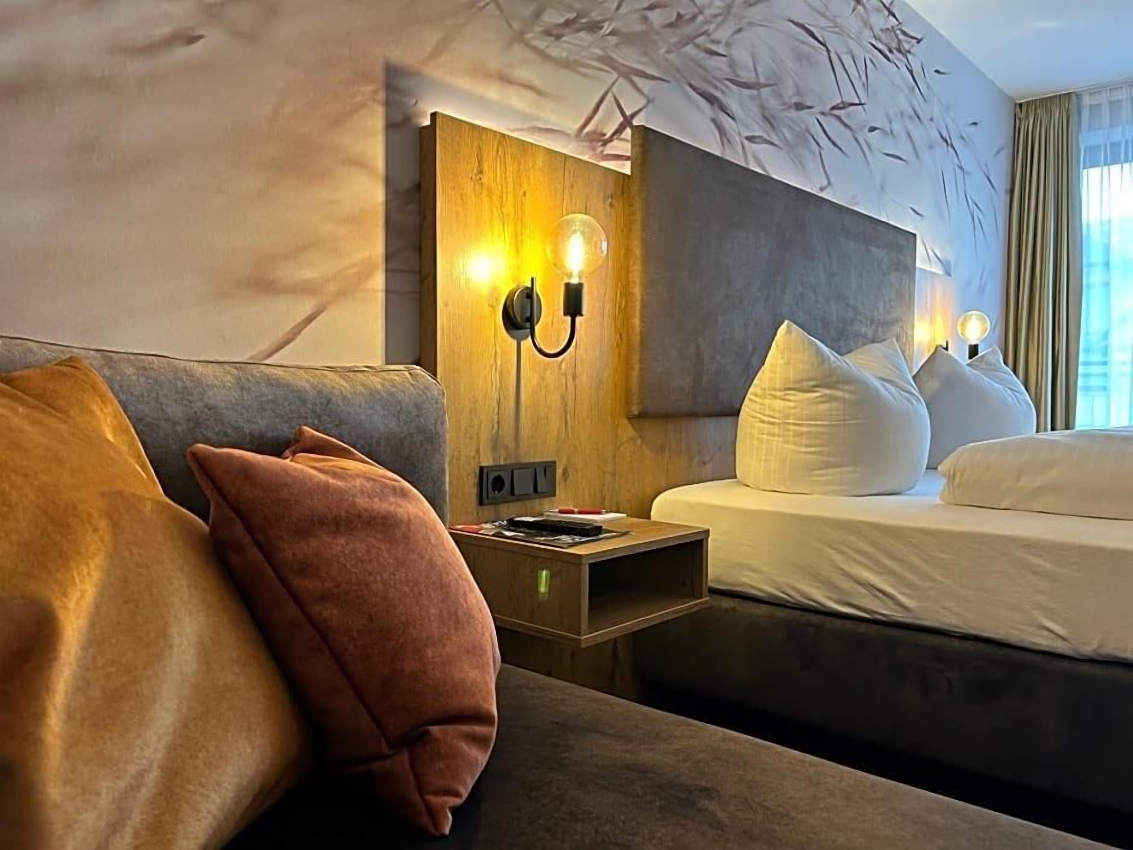A hotel room with a bed, a nightstand, and a couch. There are wall sconces with light bulbs, a wooden headboard, and a patterned wall.