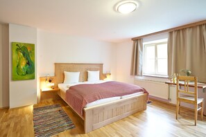 Ein Schlafzimmer mit einem hölzernen Bett, Nachttischlampen, einem Fenster mit Vorhängen, einem Schreibtisch mit Stuhl und einem farbenfrohen Teppich.