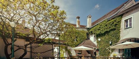 Un área de comedor al aire libre con mesas y sillas, rodeada de edificios y un árbol.