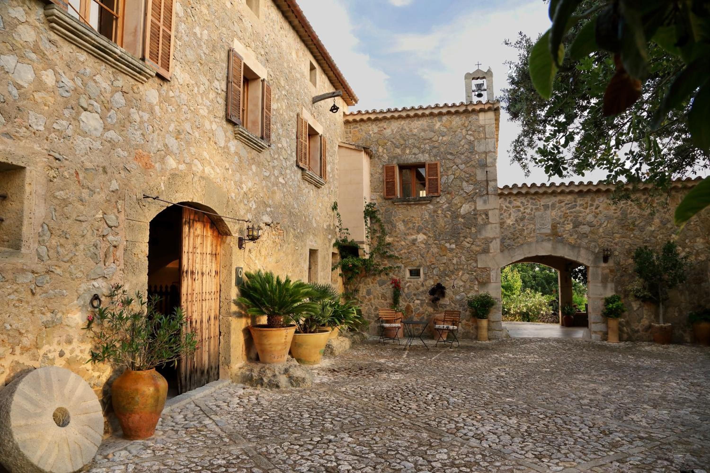 Un patio de piedra con un edificio rústico, plantas en macetas y una pequeña mesa con sillas.