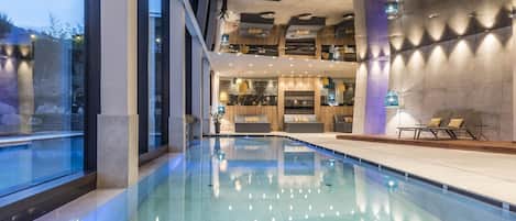 A modern indoor swimming pool with a glass barrier, lounge chairs, and a dining area with a high ceiling and pendant lighting.