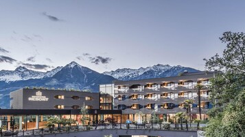 Un hotel de varios pisos con diseño moderno, con balcones y ventanas amplias, frente a un paisaje de montañas nevadas.
