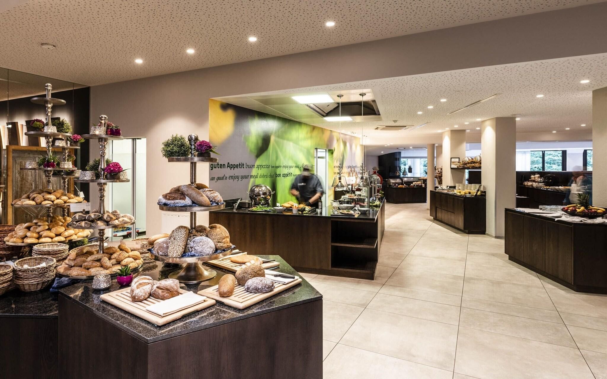 A bakery counter with various breads and pastries, a chef preparing food, and a kitchen area in the background.