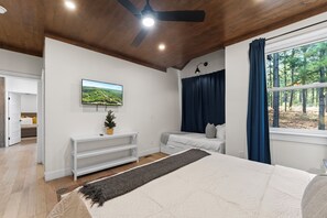 A bedroom with a bed, a television on the wall, a ceiling fan, a window with curtains, and a view of trees outside. - NEW Bigfoot Views | Sleeps 18 • Games • Playground (Broken Bow)