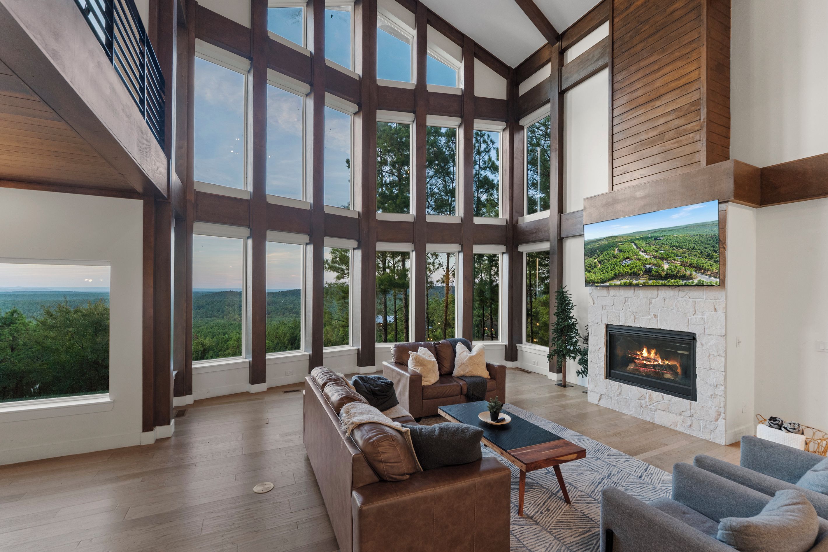 A spacious living room with a large fireplace, a sofa set, and a view of a forested landscape through large windows.
