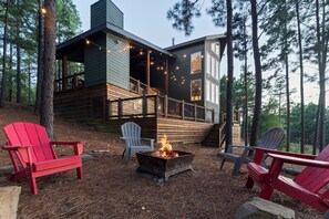 A cabin with a deck, surrounded by trees, and a fire pit with chairs. - NEW Bigfoot Views | Sleeps 18 • Games • Playground (Broken Bow)