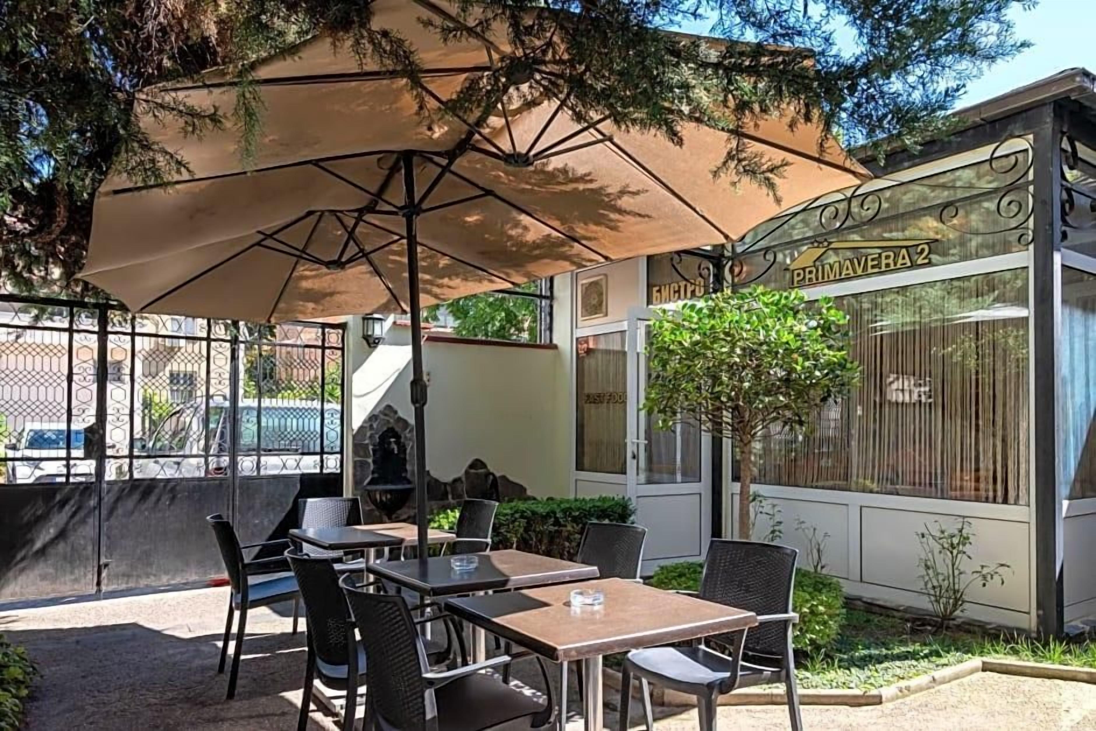 An outdoor seating area with tables and chairs, a large umbrella, and a building with a sign that reads 'PRIMAVERA 2'.