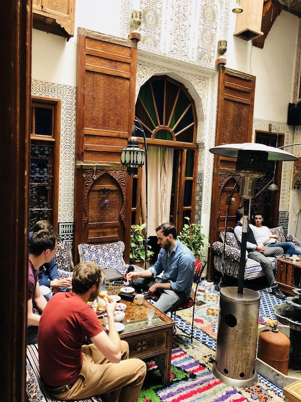 A traditional Moroccan interior with ornate wooden doors, a patterned rug, and a group of people seated around a table. - Riad Verus (Fes)
