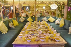 A communal dining area with a long table filled with various dishes and drinks, surrounded by sofas and a patterned wall. - Riad Verus (Fes)