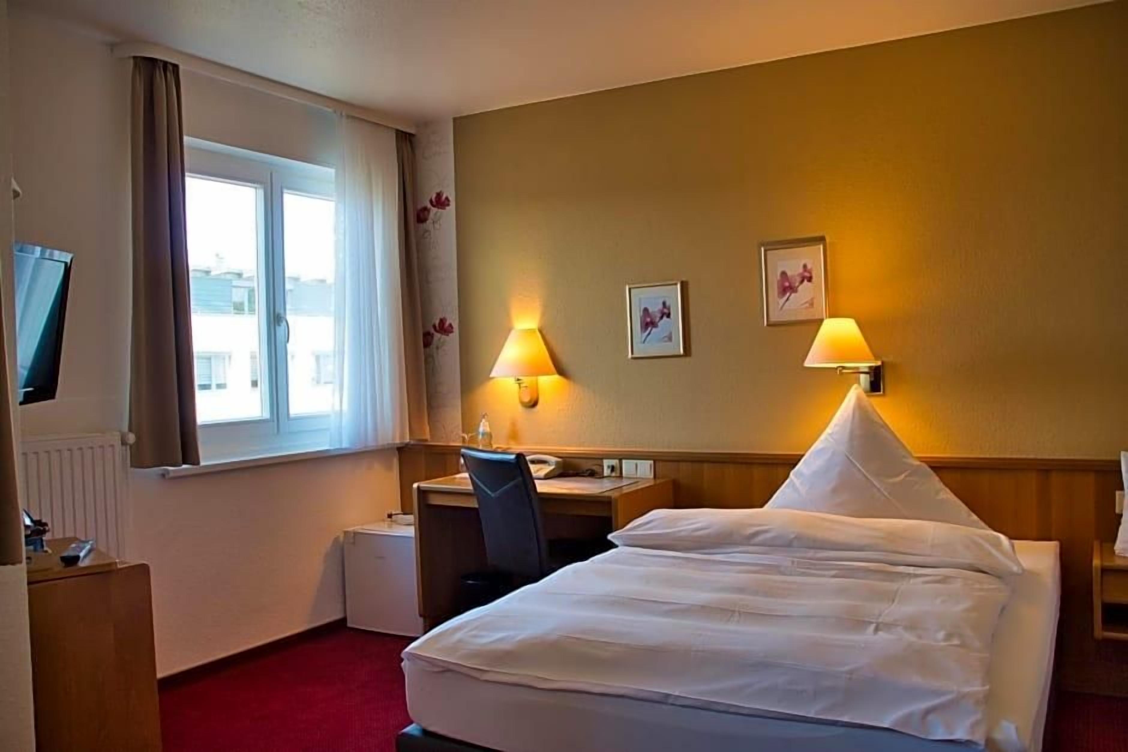 Une chambre d’hôtel avec un lit, un bureau, une chaise et un téléviseur, le tout situé à gauche.
