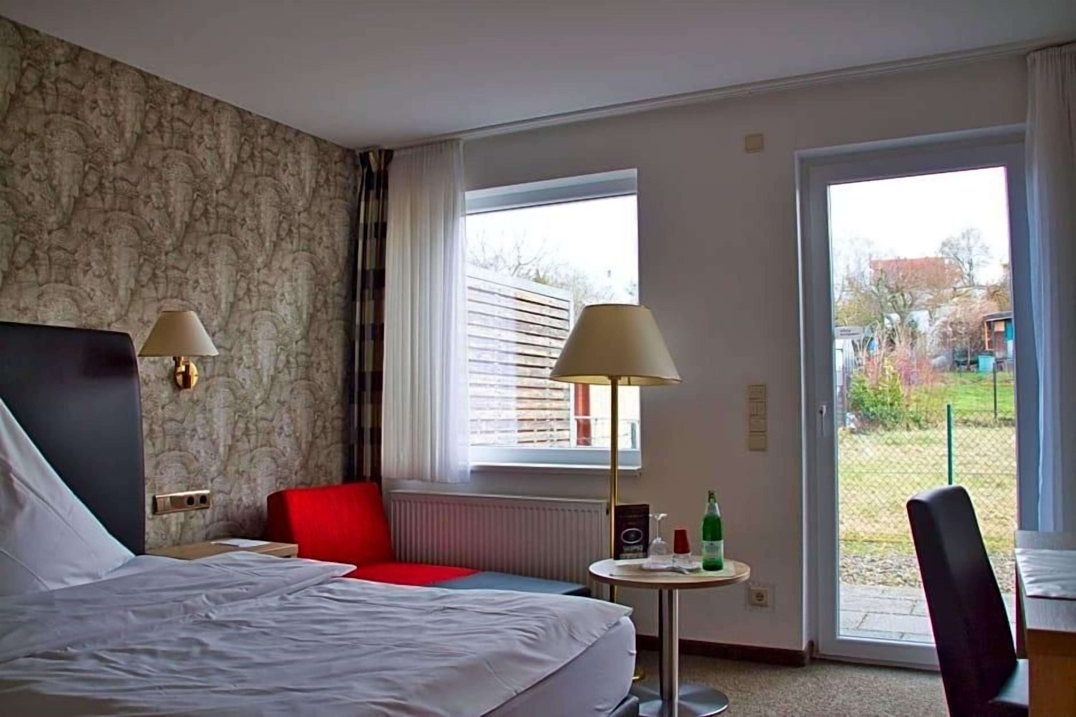 Habitación de hotel con cama, dos lámparas de noche, una silla roja, una mesita redonda con una botella y vistas a través de una ventana.