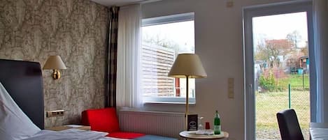 Habitación de hotel con cama, dos lámparas de noche, una silla roja, una mesita redonda con una botella y vistas a través de una ventana.