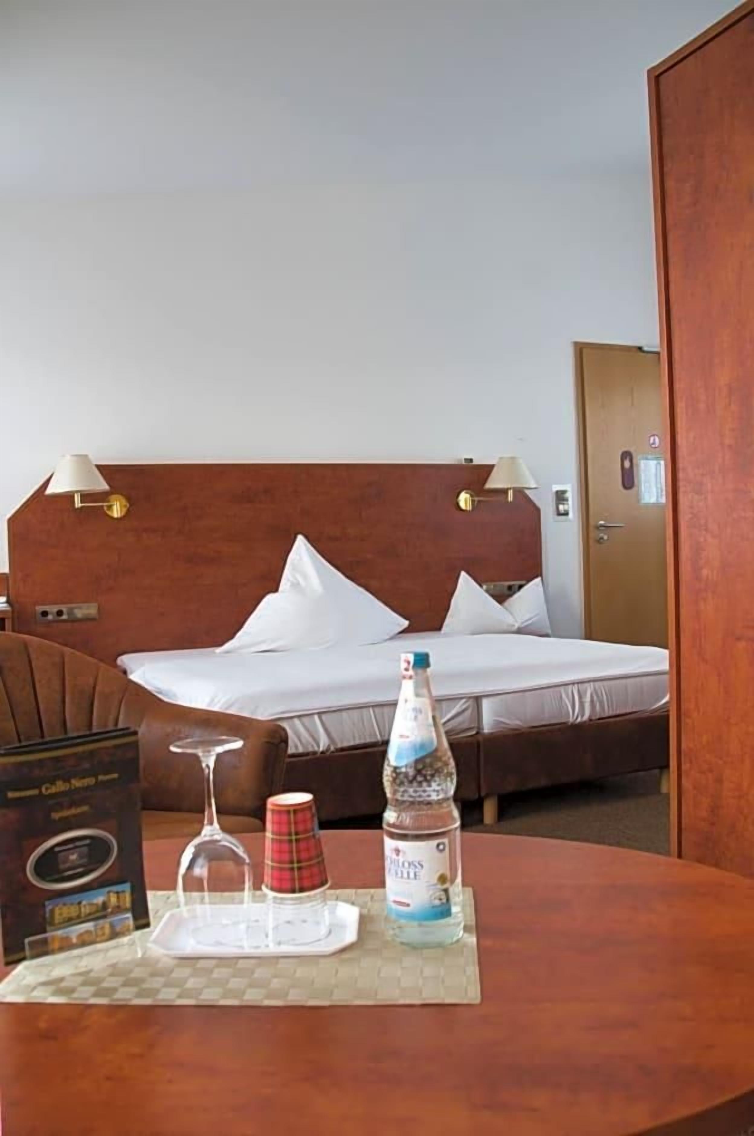 A hotel room with a bed, bedside lamps, a table with a bottle, a glass, and a box.