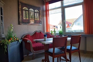 A room with a red upholstered bench, a white table, and two wooden chairs. There is a window with curtains, a painting on the wall, and a potted plant. - Quellenhof (Salzgitter)