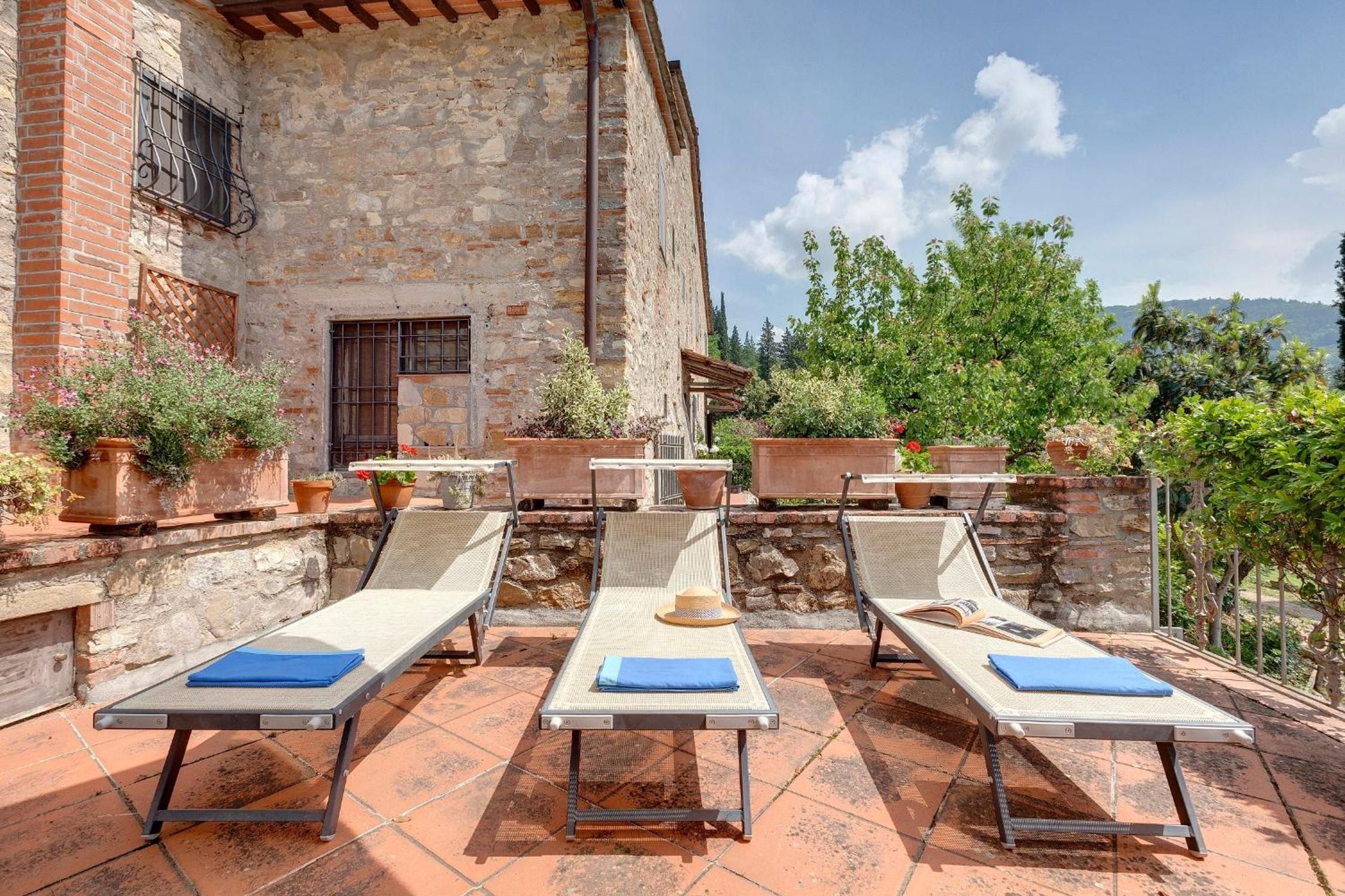 A terrace with lounge chairs and potted plants, overlooking a stone building and greenery.