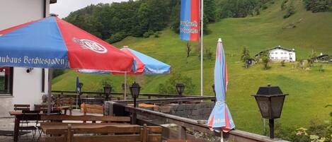 Un espace extérieur aménagé avec des tables et des chaises en bois, un drapeau et une vue sur une colline verdoyante.