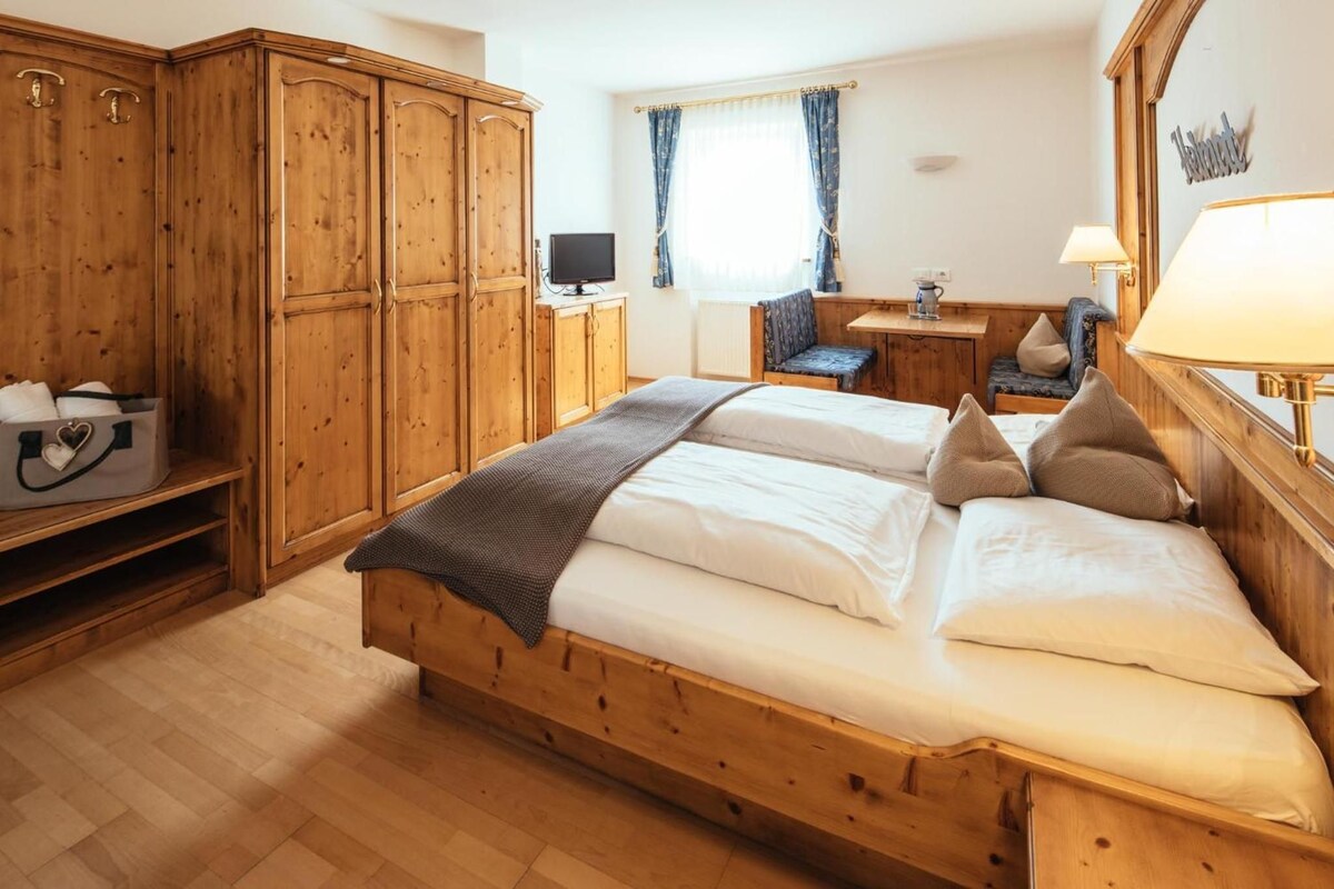 A wooden bed with pillows and a blanket, a wooden wardrobe, a television, and a window with curtains.
