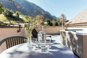 A table with a vase of flowers, a bottle, and two glasses on a balcony with a mountain view. - Tiroler Hof (San Leonardo in Passiria)