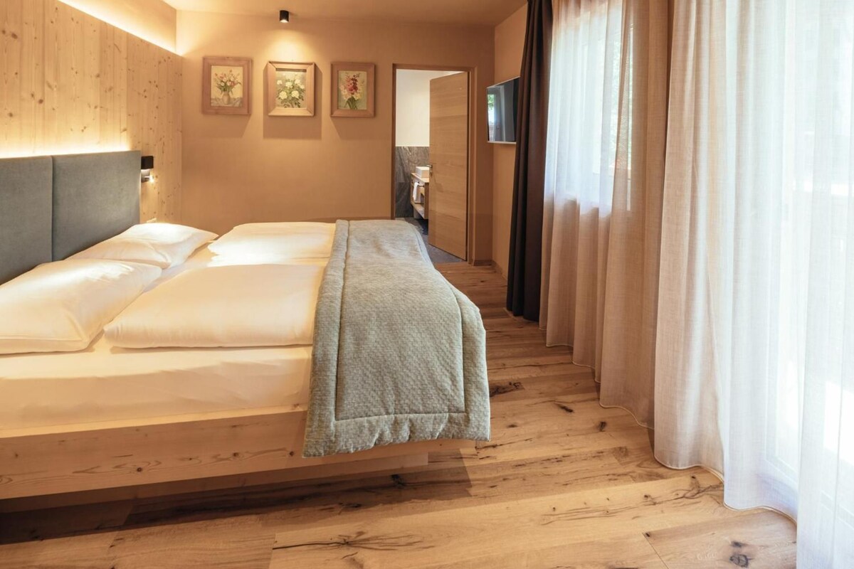 A hotel room with a bed, wooden flooring, a TV mounted on the wall, and a window with curtains.