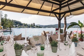An outdoor dining area with tables set for a meal, overlooking a body of water with boats and a hilly landscape in the background. - Strandhotel Sille (Maria Woerth)