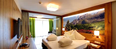 Habitación de hotel con cama, mesitas de noche, escritorio, televisor y un amplio ventanal con vista a la montaña.
