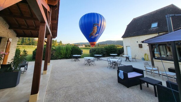 A hot air balloon in the distance, outdoor seating area with tables and chairs, and a building with a sloped roof. - L'oustal (Vezac)
