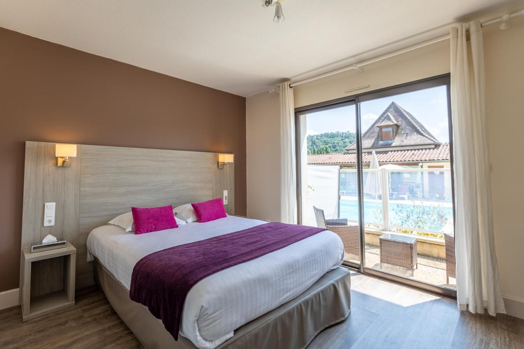 Une chambre d’hôtel avec un grand lit, une table de chevet, une fenêtre donnant sur une piscine et des bâtiments, et une porte-fenêtre coulissante.