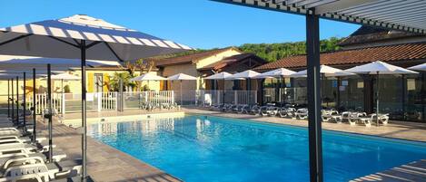A swimming pool with lounge chairs and umbrellas, surrounded by buildings with a tiled roof.