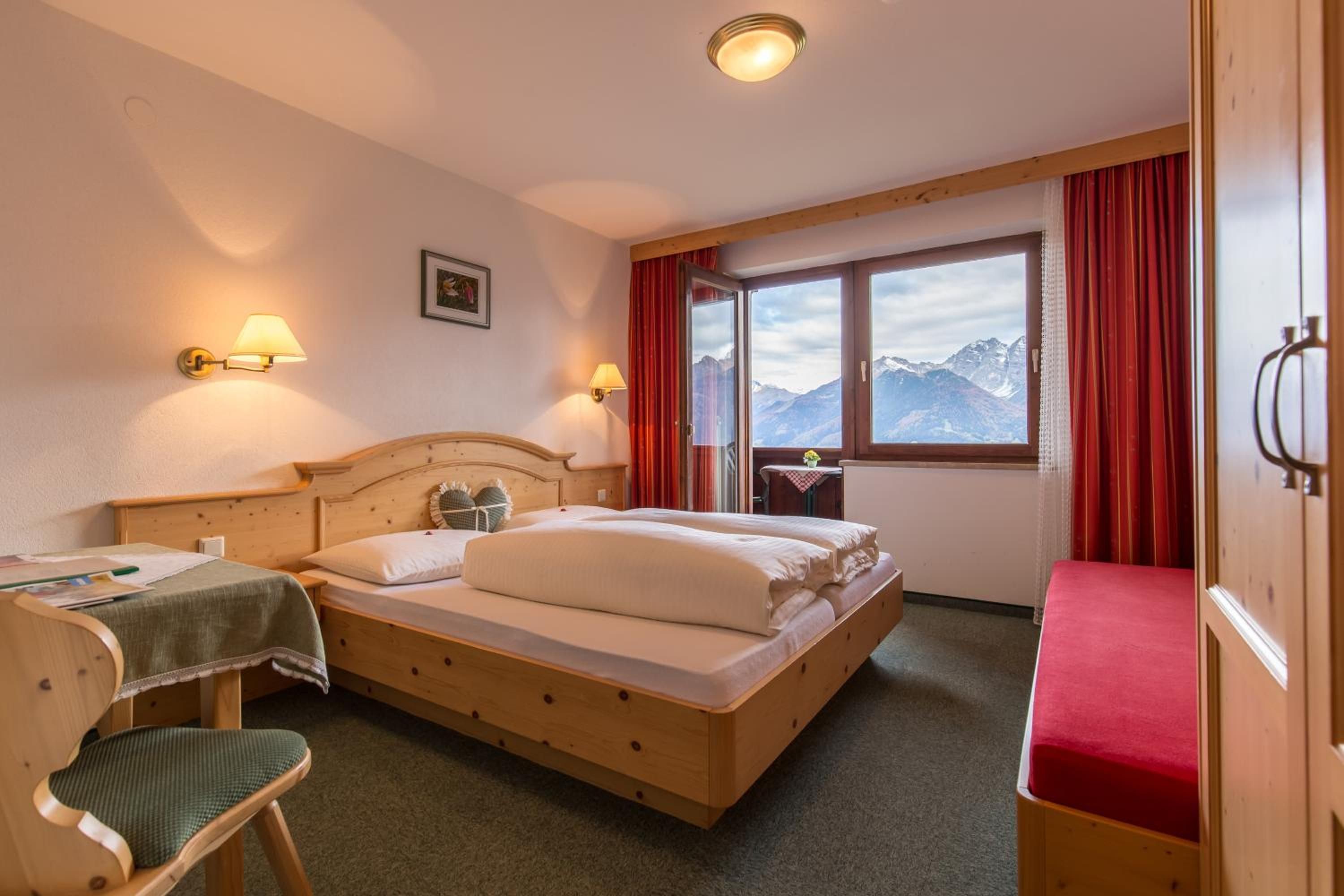 A hotel room with a wooden bed, two bedside lamps, a window with a mountain view, a small table with a vase, and a red curtain.